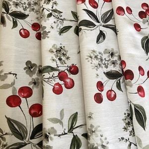 May include: A close-up of white fabric featuring a repeating pattern of red cherries, green leaves, and grey floral accents. The fabric is folded, revealing its texture and design. The cherries are a bright red against the neutral background.