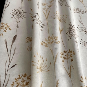 May include: Cream-coloured fabric featuring a repeating pattern of stylised brown and gold botanical illustrations. The design includes delicate stems and seed heads, creating a natural, organic aesthetic. The fabric appears slightly textured, adding depth to the design.