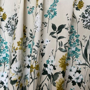 May include: A cream-coloured fabric with a floral pattern featuring white, teal, and mustard yellow flowers. The fabric has a subtle texture and a soft, flowing drape.