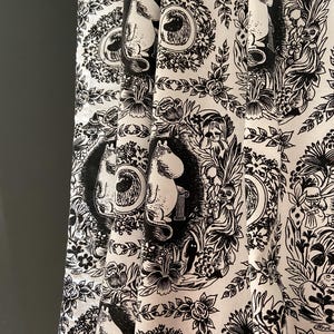 Op de afbeelding: Witte stof met een herhaald zwart patroon met cartoonfiguren, bloemmotieven en decoratieve kaders. De stof is gevouwen en toont de gedetailleerde print van de grillige illustraties. Het algemene thema is een speels, geïllustreerd ontwerp.