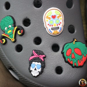 May include: A gray pair of clogs with colorful shoe charms. The charms include a green and yellow jester, a white sugar skull with colorful details, a green apple with a red dripping skull, and a black skull with pink hair and blue sunglasses.