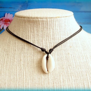 Può includere: Una collana con cordino nero con un ciondolo a conchiglia di cauri bianco. Il guscio è sospeso al cordino con un nodo. La collana è esposta su un manichino con sfondo neutro.