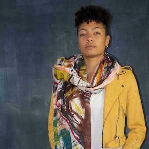 May include: A colorful scarf with a vibrant abstract design in shades of red, blue, yellow, and brown is draped over a white shirt. A yellow suede jacket with zipper details and dark blue jeans complete the look. The scarf has a fringed edge.