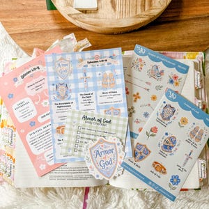 May include: A collection of religious paper study sheets and sticker sheets featuring the Armor of God theme, with illustrations of shields, swords, and floral motifs in blue, pink, and green, displayed on an open book with a wooden tray above.