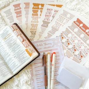 May include: An open Bible with decorative tabs, pens, and a list of Bible themes. The tabs feature various patterns and colors, including floral and geometric designs. The pens are brown, gold, and orange. The text "Bible Themes" is visible.