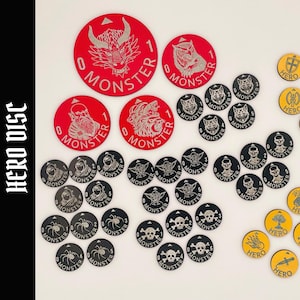 May include: A collection of circular game tokens labeled HERO DISC. The set includes large and small red tokens with monster illustrations, black tokens featuring various monster icons, and yellow tokens marked with hero symbols and the word HERO.