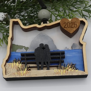 May include: Wooden ornament in the shape of Oregon, featuring a couple kissing on a bench overlooking the ocean. A heart-shaped piece with "2025" is attached. The ornament has a natural wood frame and is accented with blue, gray, and tan colors.