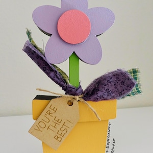 May include: A wooden flower pot with a purple flower and a pink center. The pot is yellow and has a brown tag that says "You're the best!"