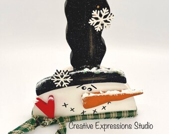 May include: A wooden snowman decoration with a black top hat, a carrot nose, and a green plaid scarf. The snowman has a red heart and snowflake accents. The text "Creative Expressions Studio" is visible.