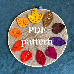 May include: A circular embroidery hoop featuring a collection of felt leaves in various autumn colors, including orange, yellow, red, and purple. The leaves are arranged around the center and attached with twine. The text "PDF pattern" is displayed in the center.