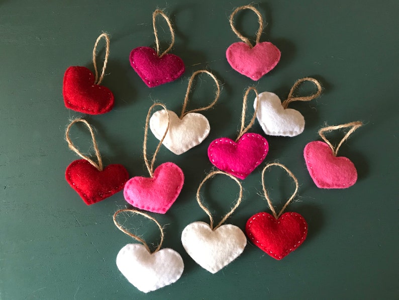 May include: A collection of twelve small heart-shaped ornaments made of felt in shades of red, pink, and white. Each heart features visible stitching along the edges and is attached to a thin, natural-colored jute twine loop for hanging.