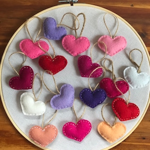 May include: A collection of small, stuffed felt hearts in various colors including red, pink, purple, white, and peach, each featuring a jute twine hanging loop and white stitched edges, arranged on a circular wooden embroidery hoop frame.