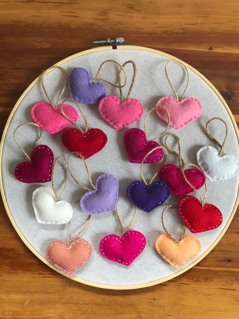 May include: A collection of small, stuffed felt hearts in various colors including red, pink, purple, white, and peach, each featuring a jute twine hanging loop and white stitched edges, arranged on a circular wooden embroidery hoop frame.