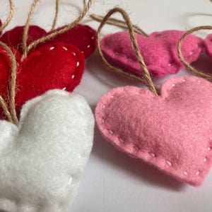 May include: A collection of heart-shaped ornaments crafted from felt in shades of red, white, and pink. Each heart features visible white stitched edges and is attached to a thin, natural-colored jute twine loop for hanging purposes.