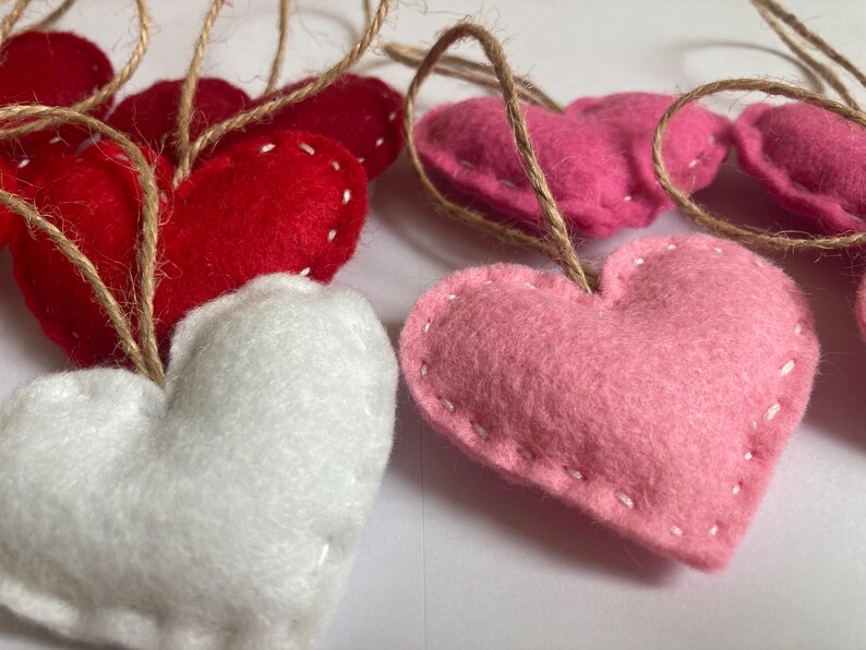 May include: A collection of heart-shaped ornaments crafted from felt in shades of red, white, and pink. Each heart features visible white stitched edges and is attached to a thin, natural-colored jute twine loop for hanging purposes.