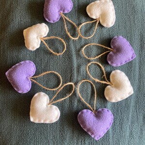 May include: A collection of seven heart-shaped ornaments made from felt in shades of lavender and white. Each heart features visible stitching along the edges and includes a small, looped jute twine string attached to the top for hanging purposes.