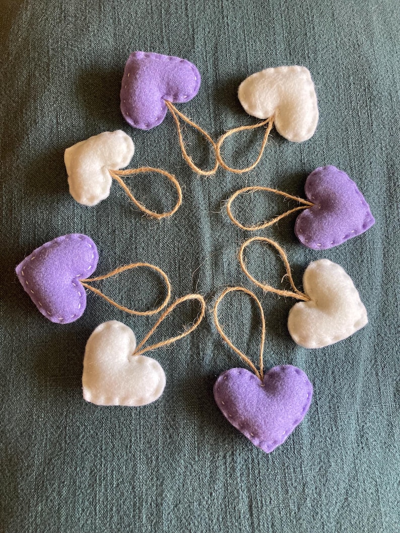 May include: A collection of seven heart-shaped ornaments made from felt in shades of lavender and white. Each heart features visible stitching along the edges and includes a small, looped jute twine string attached to the top for hanging purposes.
