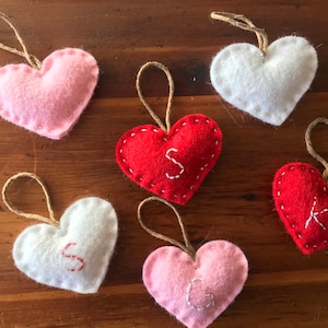 May include: Six heart-shaped felt ornaments with jute twine loops for hanging. The collection includes two white, two red, and two pink hearts. Three of the hearts feature embroidered letters: a white S on a red heart, a red S on a white heart, a white G on a pink heart, and a white K on a red heart.