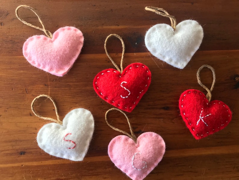 May include: Six heart-shaped felt ornaments with jute twine loops for hanging. The collection includes two white, two red, and two pink hearts. Three of the hearts feature embroidered letters: a white S on a red heart, a red S on a white heart, a white G on a pink heart, and a white K on a red heart.