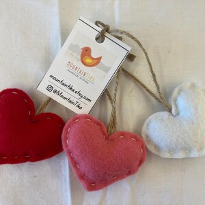 May include: Three heart-shaped felt ornaments in red, pink, and white, each featuring visible white stitching along the edges and attached to a brown twine loop. A white rectangular tag is tied to the twine, displaying a bird logo and text.