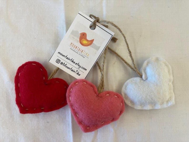 May include: Three heart-shaped felt ornaments in red, pink, and white, each featuring visible white stitching along the edges and attached to a brown twine loop. A white rectangular tag is tied to the twine, displaying a bird logo and text.