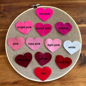 May include: A circular wooden embroidery hoop containing a tan burlap fabric background with twelve felt heart cutouts arranged in rows. Each heart is labeled with its color: hot pink, bright pink, punch pink, fuschia, pink, rose pink, light pink, white, burgundy, garnet, deep red, and red.