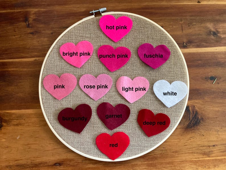 May include: A circular wooden embroidery hoop containing a tan burlap fabric background with twelve felt heart cutouts arranged in rows. Each heart is labeled with its color: hot pink, bright pink, punch pink, fuschia, pink, rose pink, light pink, white, burgundy, garnet, deep red, and red.