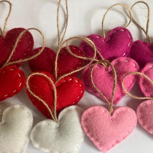 May include: A collection of heart-shaped ornaments made of felt in shades of red, pink, and white. Each heart features visible white stitching along the edges and is attached to a thin, natural-colored jute twine loop for hanging purposes.