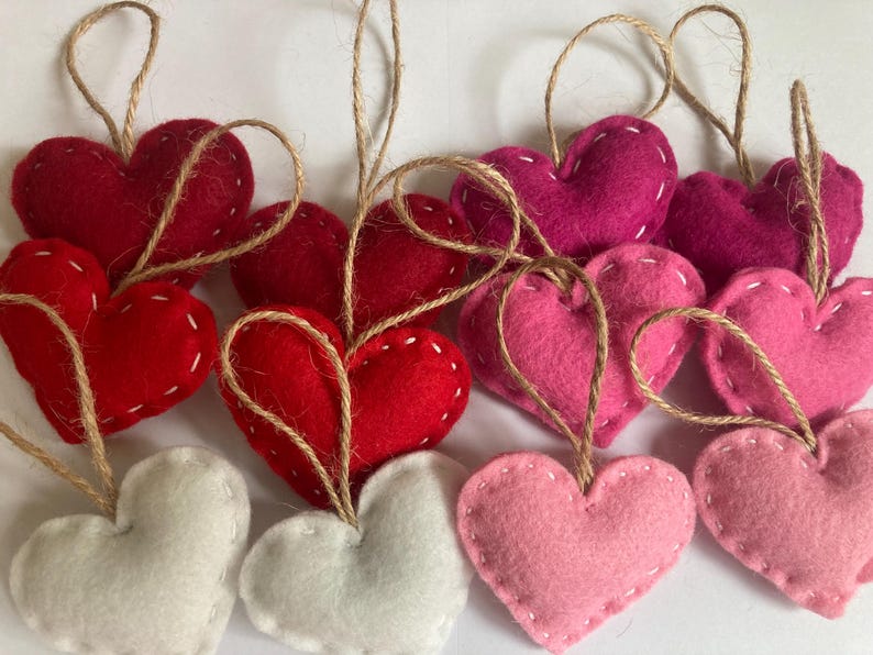 May include: A collection of heart-shaped ornaments made of felt in shades of red, pink, and white. Each heart features visible white stitching along the edges and is attached to a thin, natural-colored jute twine loop for hanging purposes.