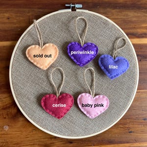 May include: Five heart-shaped felt ornaments with jute twine loops arranged on a circular embroidery hoop. The hearts are labeled with text: sold out, periwinkle, lilac, cerise, and baby pink. Each heart features a simple stitched edge.