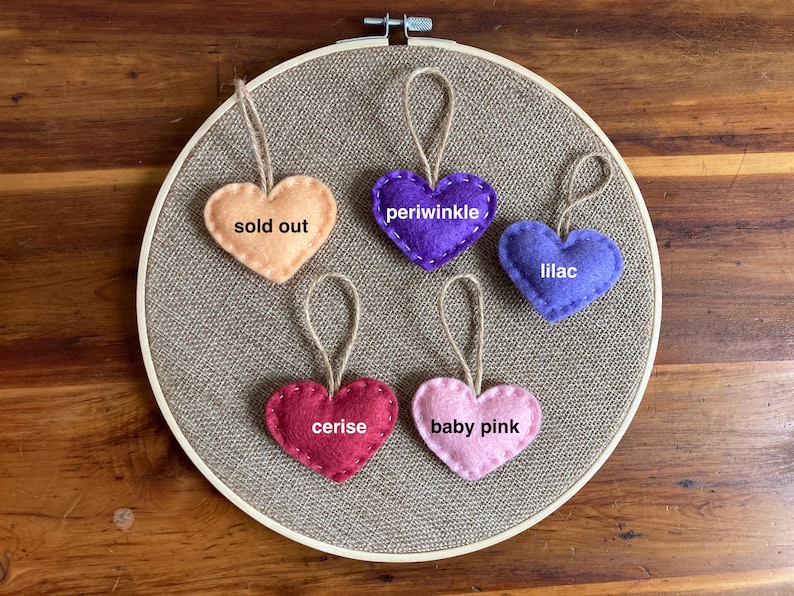 May include: Five heart-shaped felt ornaments with jute twine loops arranged on a circular embroidery hoop. The hearts are labeled with text: sold out, periwinkle, lilac, cerise, and baby pink. Each heart features a simple stitched edge.
