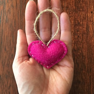 May include: A small, stuffed, heart-shaped ornament made of vibrant magenta felt. The heart features visible white stitching along its edges and includes a thin, natural-colored jute twine loop attached to the top for hanging purposes.