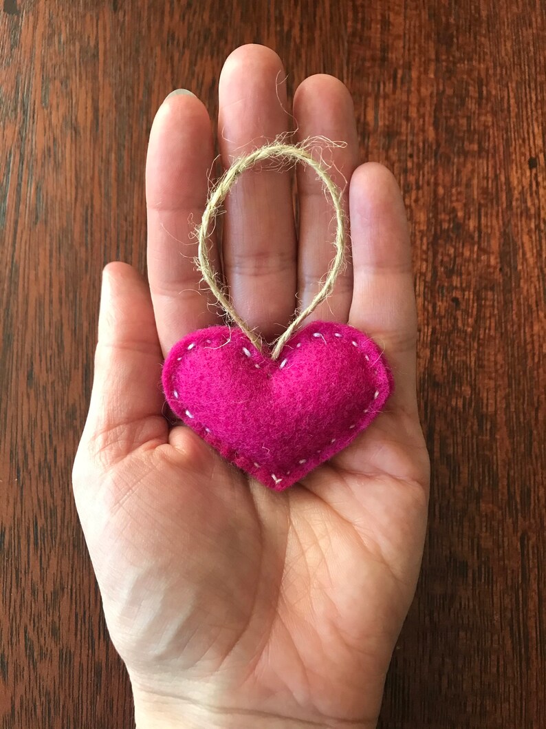 May include: A small, stuffed, heart-shaped ornament made of vibrant magenta felt. The heart features visible white stitching along its edges and includes a thin, natural-colored jute twine loop attached to the top for hanging purposes.
