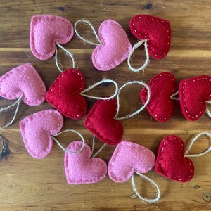 May include: A collection of twelve heart-shaped ornaments made of felt in shades of red and light pink. Each heart features visible white stitching along the edges and includes a small, attached loop of natural jute twine for hanging purposes.