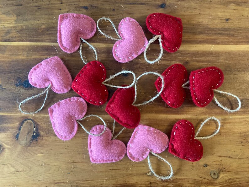 May include: A collection of twelve heart-shaped ornaments made of felt in shades of red and light pink. Each heart features visible white stitching along the edges and includes a small, attached loop of natural jute twine for hanging purposes.