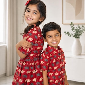 May include: Two children wearing matching red outfits with an elephant pattern. The girl is wearing a dress, and the boy is wearing a button-up shirt. Both are smiling at the camera. The outfits are made of a soft fabric.
