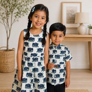 Puede incluir: Una niña con un vestido sin mangas y un niño con una camisa abotonada, ambos con un fondo blanco y un estampado de elefantes azul marino. La niña tiene trenzas y el niño lleva pantalones negros.