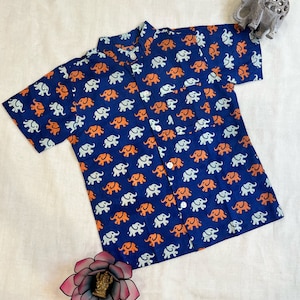 Puede incluir: Una camisa azul de manga corta con estampado de elefantes blancos y naranjas. La camisa tiene un cuello abotonado y un bolsillo en el lado izquierdo.