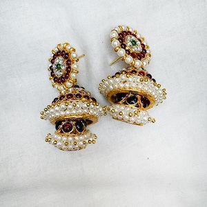 May include: A pair of gold-toned, ornate earrings. Each earring features a domed design with red and green gemstones, surrounded by small pearls. The earrings have a bell-like shape, with multiple layers of pearls and gold accents.