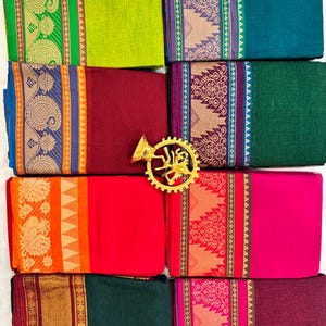 May include: An assortment of folded fabric textiles in various colors, including green, red, and teal. Each textile features a decorative border with intricate patterns in gold, orange, and purple. A gold-colored, ornate object is placed in the center.