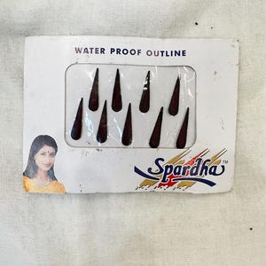 Puede incluir: Un paquete de delineadores cosméticos en forma de lágrima de color marrón oscuro. El paquete dice "WATER PROOF OUTLINE" y presenta la marca "Spardha". La imagen incluye un retrato de mujer en el lado izquierdo del paquete.