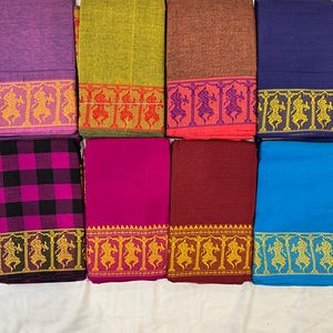 May include: Folded textiles in shades of purple, green, brown, and blue, each with a decorative border. The borders feature repeating patterns of figures in gold or red. The textiles are neatly arranged on a white surface, showcasing a variety of colours.