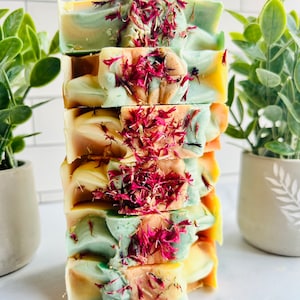 May include: A stack of handmade soap bars with a layered design in shades of green, yellow, and brown, topped with dried red flower petals. The soaps are arranged vertically, showcasing their textured surfaces and natural ingredients.