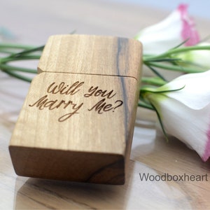 May include: A wooden ring box with the inscription "Will you marry me?" engraved on the lid. The box is closed and sitting on a wooden surface with pink and white flowers.