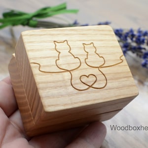 Könnte beinhalten: Eine Holzkiste für Ringe mit einem geschnitzten Design aus zwei Katzen, deren Schwänze ineinander verschlungen sind und ein Herz bilden. Die Box ist aus hellem Holz gefertigt und hat eine natürliche Oberfläche.