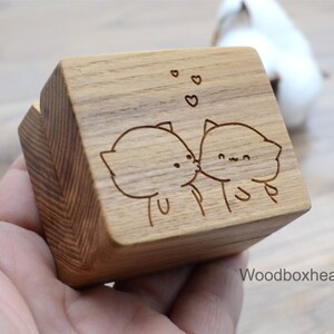 Puede incluir: Una caja de madera con un diseño grabado con láser de dos gatos de dibujos animados besándose. La caja está hecha de madera marrón claro y tiene una veta de madera natural. Los gatos están dibujados en un estilo simple con ojos grandes y bocas pequeñas. Por encima de los gatos hay tres pequeños corazones.