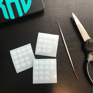 May include: Three white vinyl sticker sheets with text, a metal tool, and scissors on a dark surface. The stickers have the text "EMP" and "1PD" repeated. A black phone case with a teal design is in the background.