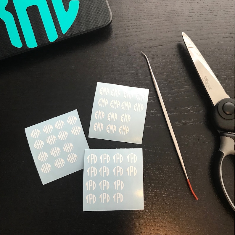 May include: Three white vinyl sticker sheets with text, a metal tool, and scissors on a dark surface. The stickers have the text "EMP" and "1PD" repeated. A black phone case with a teal design is in the background.