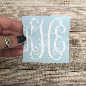May include: A white vinyl sticker with a monogram of the letters KHE in a cursive script font on a light blue background.