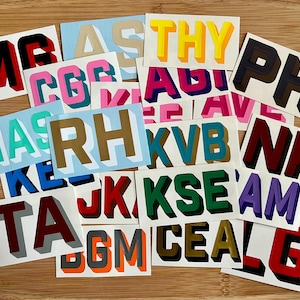 May include: A collection of colorful, 3D-style letter stickers arranged on a wooden surface. The stickers feature various letter combinations in bold, vibrant colors with shadow effects, creating a layered appearance. The letters are in different fonts.